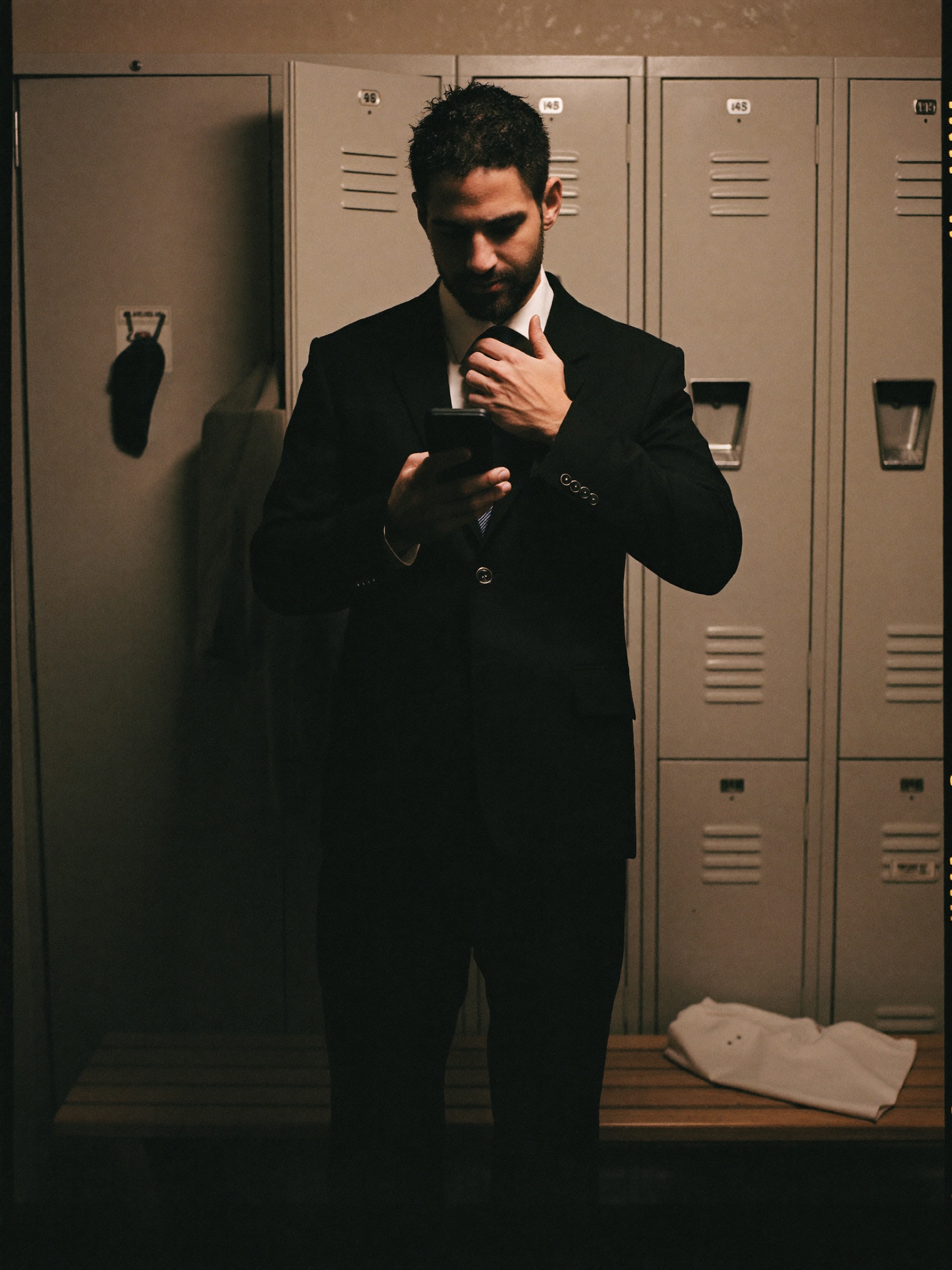A man in a suit loosening his tie in a gym locker room, reading his phone